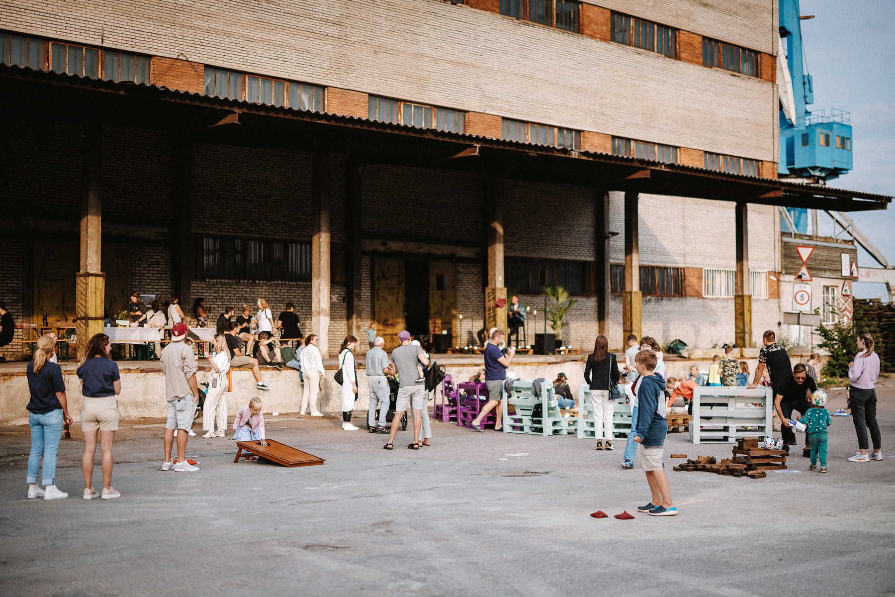 Pikalauapiknik Hundipeal. Vana laohoone esine pika lauaga, laua ääres on erinevas vanuses inimesi nautimas seltskonda, laval esinevat kitarriga muusikut ja hilissuvist päikest. Lapsed mängivad murumänge.