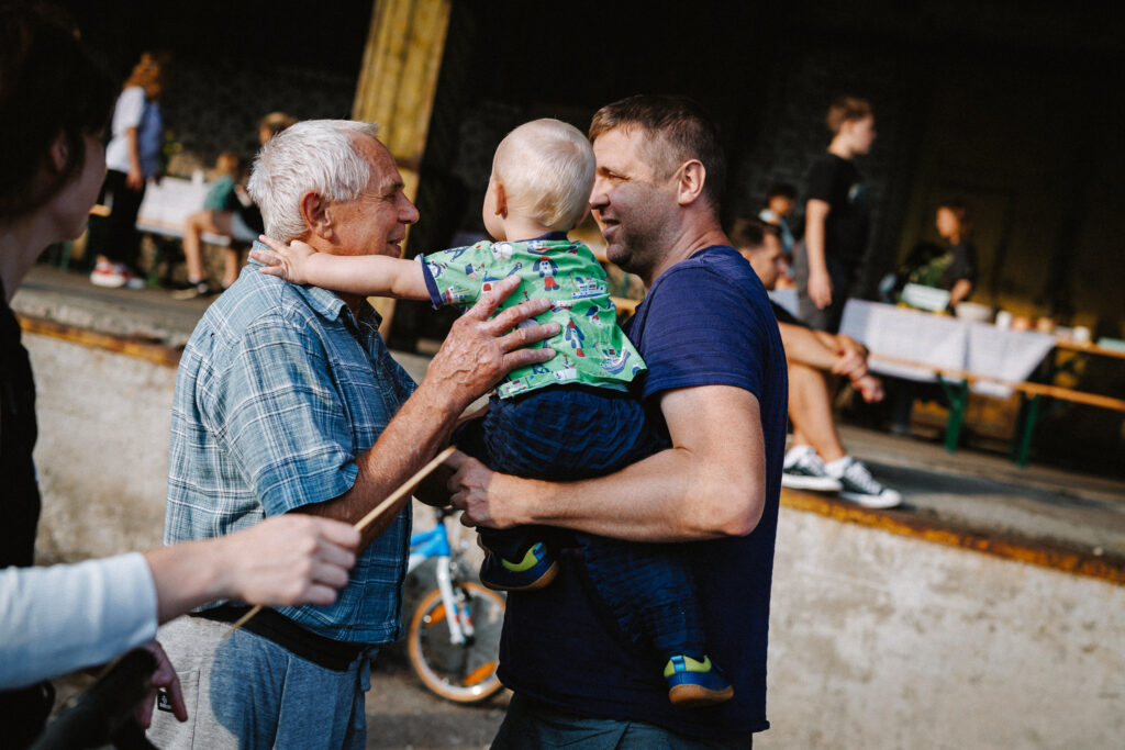 Foto Hundipea kogukonda kaasavast pikalauapiknikust suve lõpus. Pildil isa, noorem mees, ulatamas sülest pisikest last üle vanaisale, vanemale mehele. Taustal on vana laohoone, rõõmsad inimesed, piknikumelu. Õues on suvine ilm.