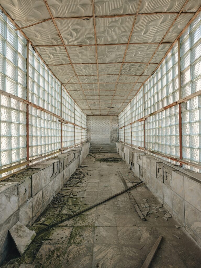 Open House Tallinn tuur Hundipeal külas. Pildil pikk koridor, mida ääristavad klaasruutudest aknad. Põrandal katkised keraamilised plaadid. Sild kahe hoone vahel. Helerohekas valgus kumab läbi akende.