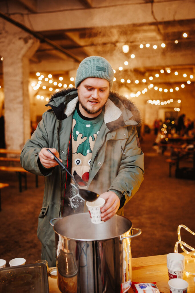 Roheline jõululaat Hundipeal. Rohelises põhjapõdraga jõulukampsunis noormees tõstab kulbiga suurest kastrulist topsikusse kuuma jõulujooki. Taustal kollased tuled ja laadamelu.