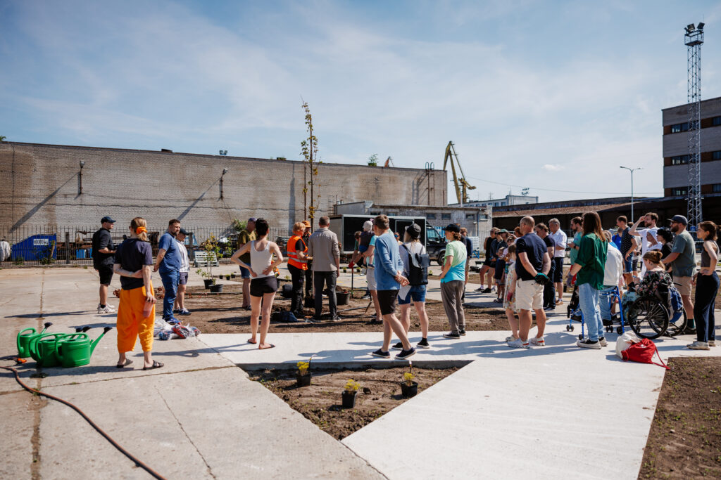 Minimetsa istutamine vana betoonplatsi vahele. Paarkümment inimest valmistumas istutamistöödeks, mullalappidel on potid taimedega, õues on ilus päikeseline ilm.