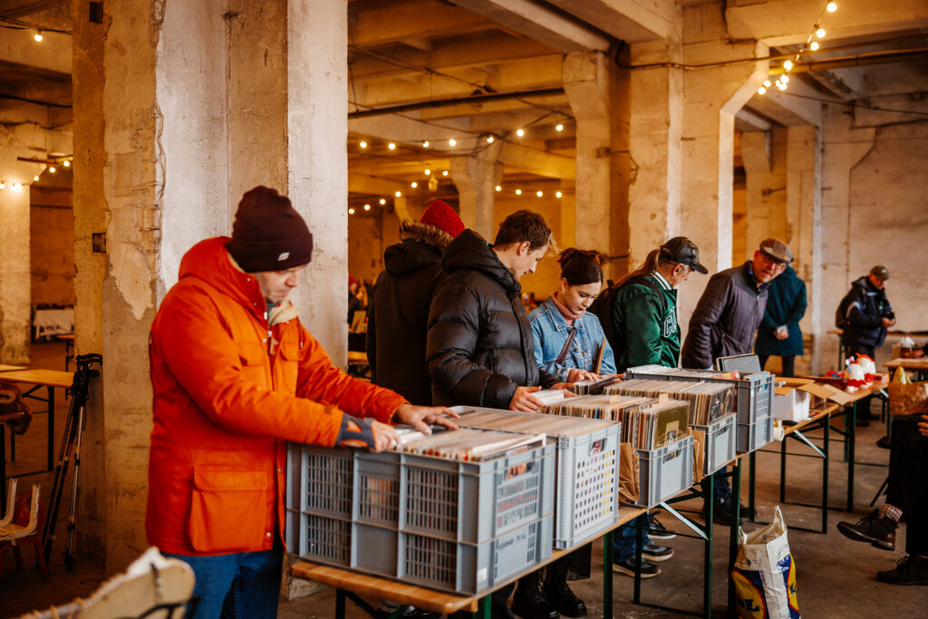 Vana kivist seintega ja kõrgete lagedega laoruum, mille teevad hubaseks soe valguskett. Pildil on 7 erinevas eas värvilistes jopedes inimest, kes sobravad vinüülikastides. See on kevadlaat.