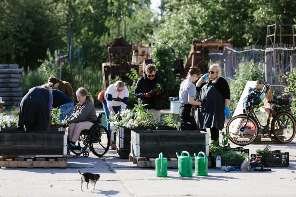 Fotol on näha 8 inimest toimetamas puidust kasvukastide ümber, istutamas taimi. Tausta puud ja rohelus, esiplaanil kivipind. Ilm on päikeseline.